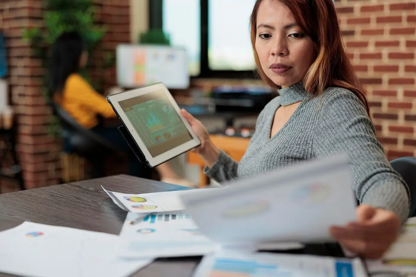 Азиатская менеджер держит планшетный компьютер и проверяет управленческую статистику, работая над стратегией компании в офисе стартапа. Деловая женщина анализирует бумаги с графиком, планируя проект партнёрства.